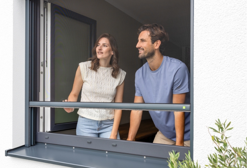 Zwei Personen stehen im Haus am Fenster und schauen durch das quasi unsichtbare Insektenschutz-Gewebe des Rollos.